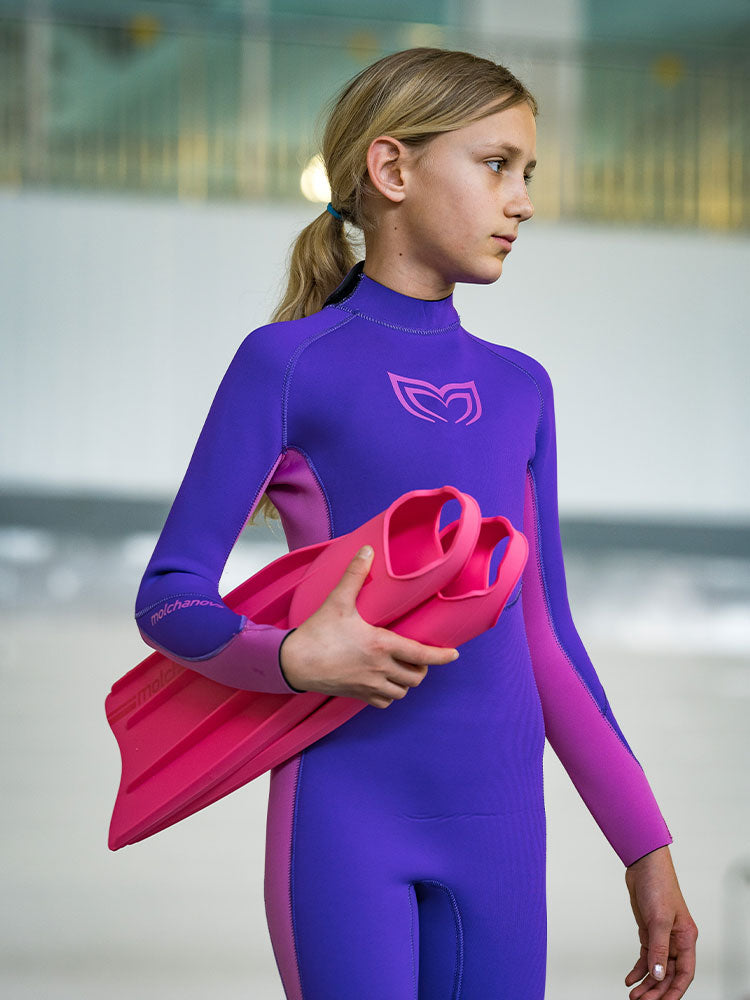 A girl wearing a purple and pink Molchanovs kids freediving wetsuit and holding a pair of fins