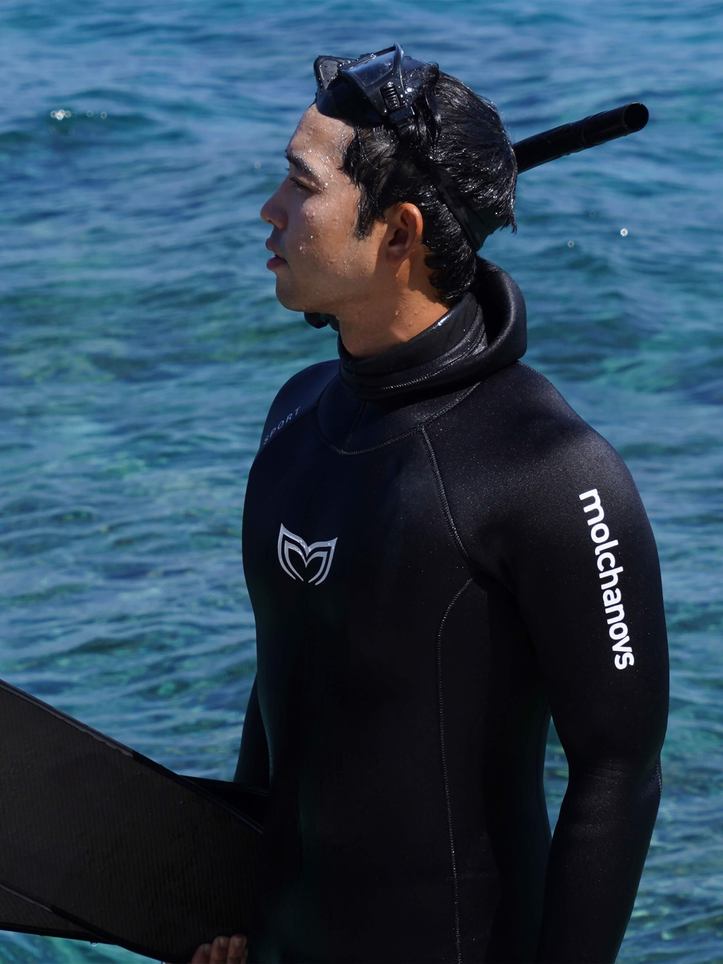 A male diver standing infront of the water wearing a black, double lined wetsuit