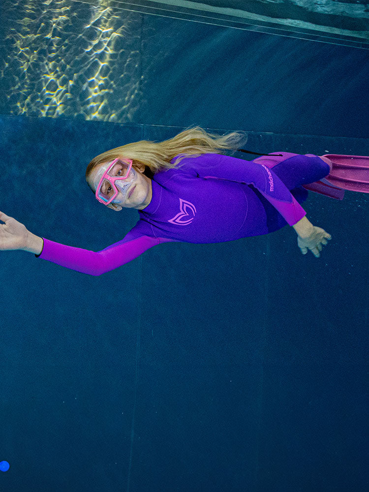 A girl swimming underwater wearing a Molchanovs wetsuit along with mask and fins