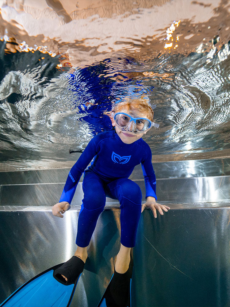 A photo from underwater of a boy wearing a wetsuit sitting on a pool step and looking down towards the camera