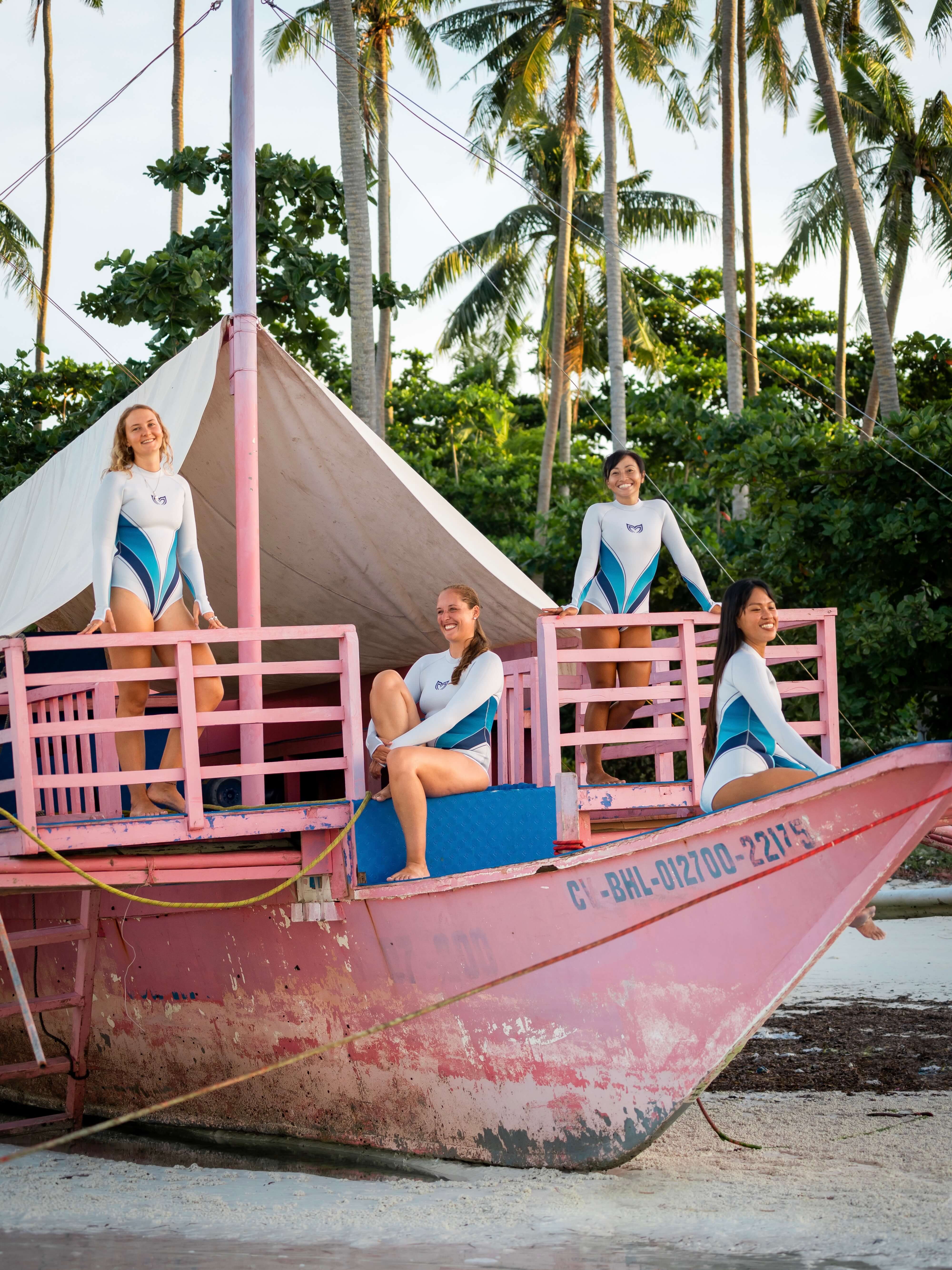 Four women in Molchanovs Yanita springsuits posing on a traditional pink wooden boat on a tropical beach